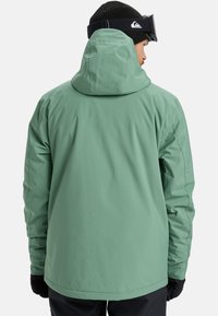 Grüne wasserdichte Jacke mit hohem Kragen, verstellbarem Kapuze und schwarzen Handschuhen. Glatte Textur, entworfen für Outdoor-Aktivitäten.