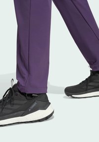 Pantaloni sportivi viola abbinati a scarpe Terrex nere con superficie testurizzata, lacci e suola bianca, fotografati da un angolo laterale basso.