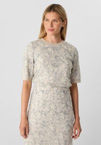 Robe beige avec broderies florales blanches et bleues, manches courtes, encolure ronde, tissu légèrement texturé, silhouette ajustée.