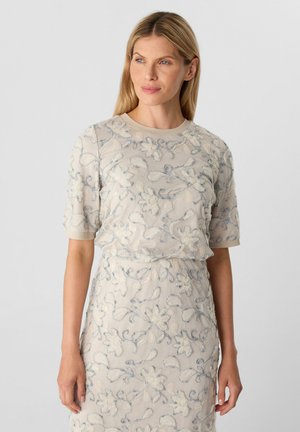 Robe beige avec broderies florales blanches et bleues, manches courtes, encolure ronde, tissu légèrement texturé, silhouette ajustée.