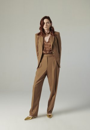 Completo marrone a righe con giacca strutturata e pantaloni morbidi, abbinato a un top marrone con fantasia. Ballerine dorate a punta e collana a catena.