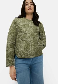 Olivengrøn quiltet jakke med hvide paisley-mønstre, der har rund halsudskæring, trykknaplukning og sidelommer, kombineret med blå jeans.