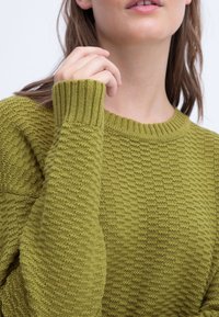 Gehaakte trui in olijfgroen; beschikt over een textuurweefsel, geribbelde manchetten en een ronde halslijn. Close-up van het schouder- en handdetail.