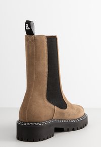 Bruine suède enkelboots met zwarte elastische zijpanelen, dikke zwarte rubberen zolen en een treklipje aan de achterkant. Accentueert met maatwerk stiksels.