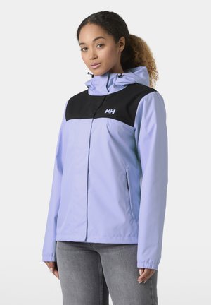 Kobieta w jasnofioletowej i czarnej kurtce z kapturem z logo Helly Hansen, w szarych dżinsach, stojąca na białym tle.