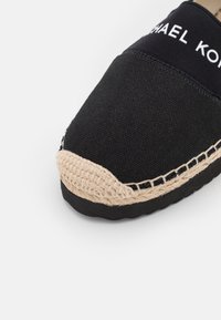 Espadrille en tissu noir avec bout tressé beige, semelle noire épaisse et logo "MICHAEL KORS" sur le bord supérieur.