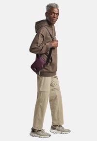 Paarse schoudertas met een ritsluiting, gedragen over een bruine hoodie en beige broek. Sneakers zijn grijs met gestructureerde accenten.