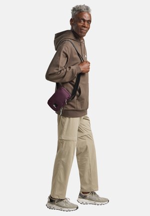 Sac en bandoulière violet avec une fermeture éclair, porté sur un sweat à capuche marron et un pantalon beige. Les baskets sont grises avec des accents texturés.