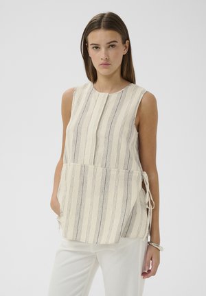 Kobieta w bezowym topie bez rękawów z pionowymi szarymi paskami i bocznymi troczkami, w połączeniu z białymi spodniami na jednolitym tle.