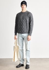 Pelēks džemperis ar pīts rakstu, gaiši zili džinsi, melnas slip-on kurpes, rokās turot bēša krāsas iepirkumu somu, galvā melna cepure ar logo.