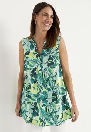 Uśmiechnięta kobieta w bezrękawnikowej tunice z zielonym i żółtym tropikalnym wzorem liści, stojąca na jednolitym białym tle.