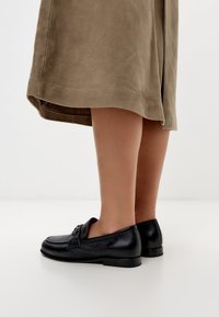 Černé kožené mokasíny s texturovaným povrchem, zdobené malou dekorativní sponou na přední straně a nízkým plochým podpatkem.