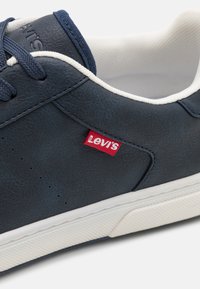 Mornarski kožni tenisice s bijelim gumiranim đonom. Imaju crveni Levi's logo tag, teksturirani gornji dio i tonirane vezice. Perforacije ukrašavaju bočne strane.