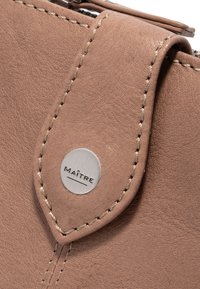 Braunes Lederaccessoire mit sichtbaren Nähten, einer abgerundeten Klappe und einem metallischen Logo-Button mit der Aufschrift "MAÎTRE". Die Struktur der Oberfläche ist deutlich erkennbar.