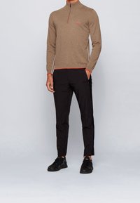 Man i en brun zip-up-tröja med röda detaljer, svarta slim-fit-byxor och svarta sneakers står mot en enkel ljus bakgrund.