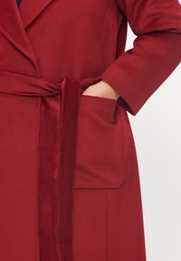 Robe de tecido vermelho e suave com um cinto e detalhes de bolso. A textura parece lisa e o design apresenta um colarinho de chal e mangas ajustadas.