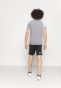 Grå treningsskjorte med blå aksenter, paret med sorte shorts med hvite chevron-mønstre. Hvite sko med oransje såler.