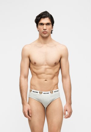 Męski mężczyzna o ciemnych włosach noszący jasnoszare slipy Moschino z białym pasem w talii z ilustracjami misiów i logo.