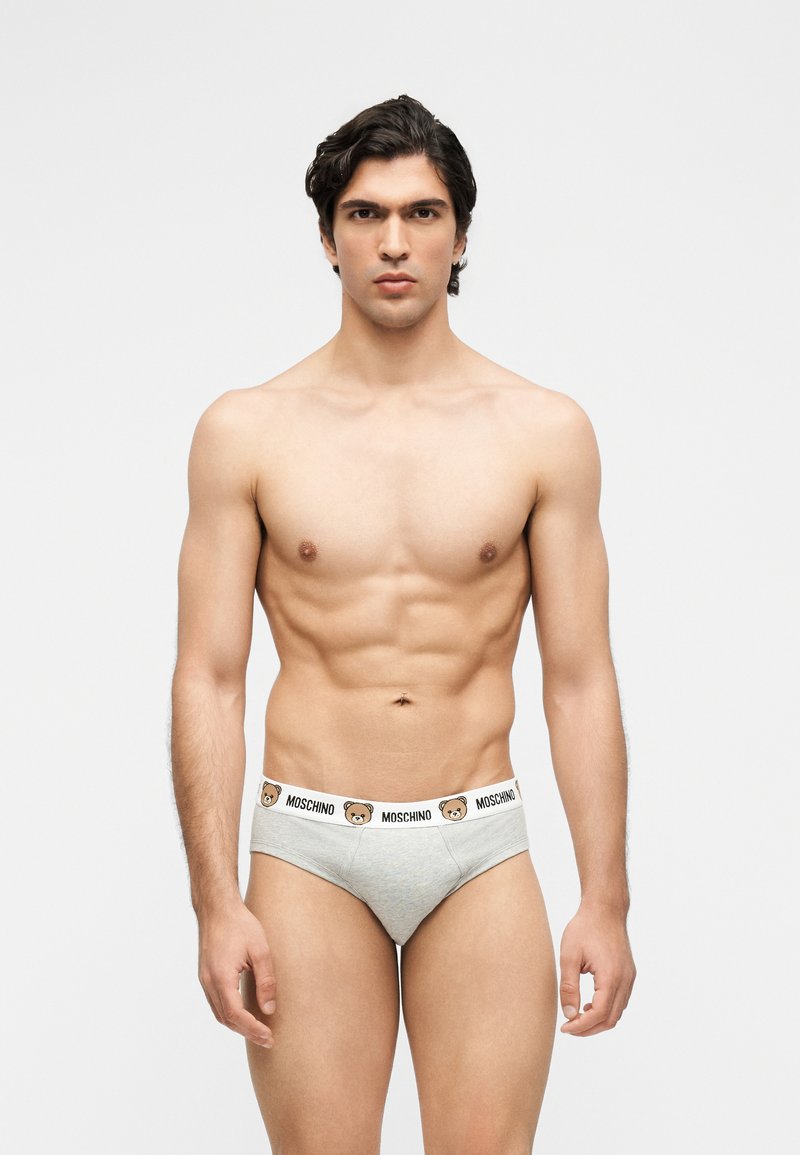 Μυώδης άνδρας με σκούρα μαλλιά φοράει ανοιχτό γκρι εσώρουχα Moschino με λευκή ζώνη μέσης, διακοσμημένη με εικόνες αρκούδων και λογότυπο.