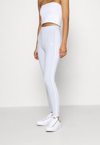 Hellgraue Leggings mit einem figurbetonten Design, die drei weiße horizontale Streifen an den Seiten und ein besticktes Logo an der Hüfte aufweisen.