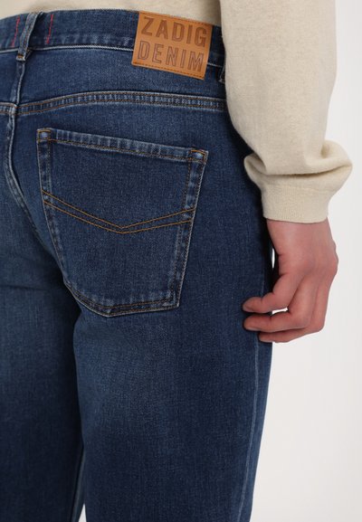 Jeans en denim bleu foncé avec une texture lisse. Comprend une étiquette en cuir marron portant l'inscription "ZADIG DENIM" et des surpiqûres contrastées sur les poches.