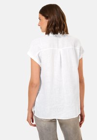 Camicia bianca di lino con maniche corte arrotolate, yoke posteriore e una texture sottile. Abbinata a pantaloni grigi chiari, l'attenzione è rivolta al design della camicia.