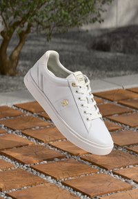 Sapata de couro branca com uma textura suave, bico arredondado e design de atacadores. Apresenta um detalhe de logotipo dourado na lateral.