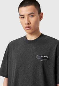 T-shirt en coton gris foncé avec un col rond et des manches courtes. Le logo affiché est partiellement obscurci par un texte en japonais et la mention "LONDRES".
