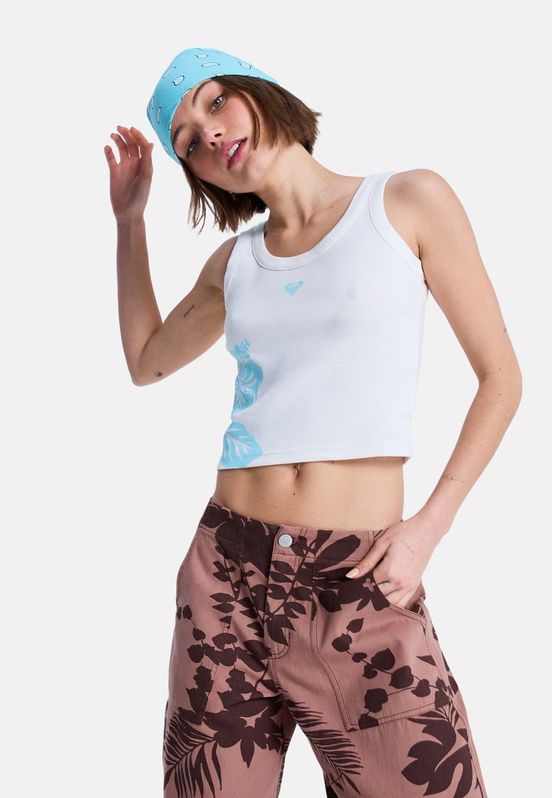 Joven mujer con una camiseta blanca corta con estampado azul, pantalones marrones con flores y un pañuelo azul posa con una mano levantada y la otra en el bolsillo.