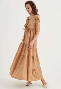 Maxi-Kleid aus beigefarbenem Crinkle-Stoff mit rundem Halsausschnitt, kurzen Ärmeln und Schleifendetail vorne. Ausgestellte Schichten sorgen für Struktur.