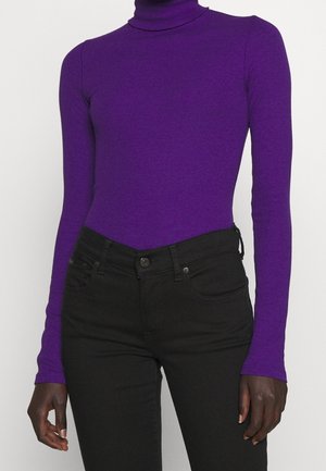 Personne portant un pull à col roulé violet ajusté et un jean noir taille haute, debout devant un fond clair uni.