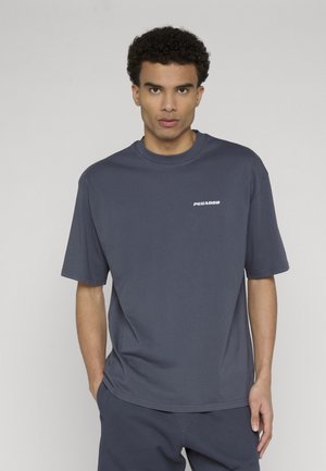 Młody mężczyzna o kręconych ciemnych włosach, ubrany w luźną granatową koszulkę z napisem "PEGADOR" na piersi, stojący z jedną ręką w kieszeni.