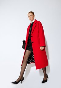 Abrigo de lana rojo con corte holgado, grandes botones y bolsillos laterales, superpuesto a un vestido negro brillante con lunares blancos y medias negras transparentes.