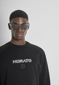 Sudadera negra con el logo "MORATO" en blanco, que presenta un emblema de tigre texturizado. Gafas de sol transparentes con diseño angular, llevadas por un modelo.