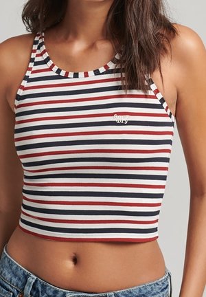 Woman wearing a sleeveless, cropped tank top with horizontal red, white, and navy stripes and denim jeans.