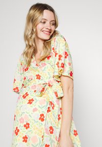 Vestido floral em tecido leve e suave, com um fundo amarelo pastel, flores vermelhas e rosas, mangas curtas e soltas, e uma cintura atada.
