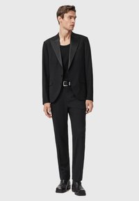 Uomo che indossa un blazer nero con rever in satin, cintura nera, pantaloni neri, stivali neri e una camicia nera, in piedi contro uno sfondo semplice.
