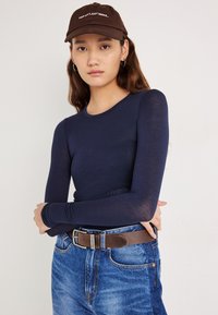 Top de manga larga color azul marino hecho de una tela ligera, combinado con vaqueros azules y una gorra marrón con texto. Cinturón marrón con herrajes plateados.