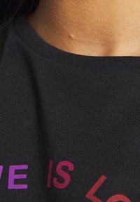 Camiseta de algodón negra con mangas cortas que presenta el texto "EL AMOR ES" en colores degradados que van del púrpura al rojo. El cuello es redondeado.