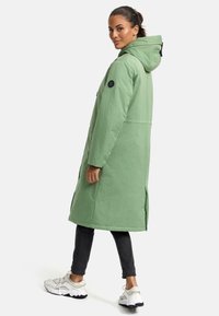 Long green puffer coat with a hood, side slits, and black logo patch, paired with dark jeans and light-colored sneakers.