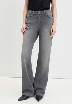 Person trägt hoch taillierte, weit geschnittene graue Jeans, schwarze spitze Schuhe und ein schwarzes kurzärmeliges Oberteil, steht mit entspannten Händen.