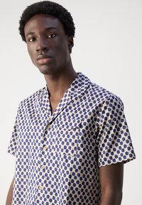 Camisa de manga corta en beige claro con un patrón de puntos azules, que cuenta con un bolsillo, cuello con solapa y botones en la parte delantera. Textura de tela suave.