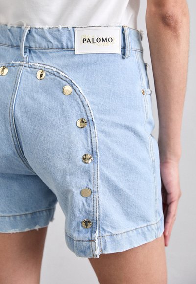 Shorts en denim bleu clair avec un ourlet effiloché, ornés de boutons dorés métalliques sur le côté et d'une étiquette PALOMO sur la taille.