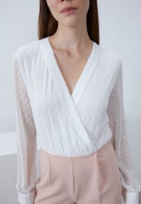 Blusa blanca de tela texturizada y con puntos, de mangas largas, combinada con pantalones rosa claro. Presenta un diseño envolvente y escote en V.