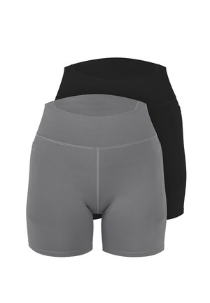 Frau steht barfuß, trägt ein graues sportliches Cropped-Top mit weißen Trägern und passende hoch taillierte Biker-Shorts vor schlichtem Hintergrund.