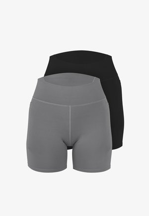Frau steht barfuß, trägt ein graues sportliches Cropped-Top mit weißen Trägern und passende hoch taillierte Biker-Shorts vor schlichtem Hintergrund.