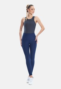 Giovane donna con top blu navy senza maniche e leggings, con scarpe da ginnastica bianche, in piedi con una mano sul fianco, guarda verso il basso sorridendo.