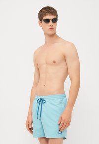 Pantaloni de baie de culoare albastră deschisă, cu talie elastică, șnur de ajustare și o mică etichetă cu logo. Purtați cu ochelari de soare negri. Fără alte articole de îmbrăcăminte.