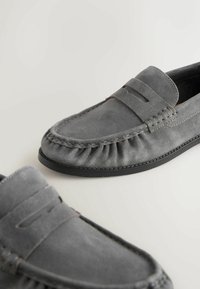 Mocassins gris en daim, sans lacets, avec couture style mocassin et semelles en caoutchouc noir sur un fond clair.