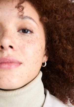 Close-up of a woman with curly reddish-brown hair wearing a cream turtleneck. Notable sparkling hoop earrings. Visible freckles on face.